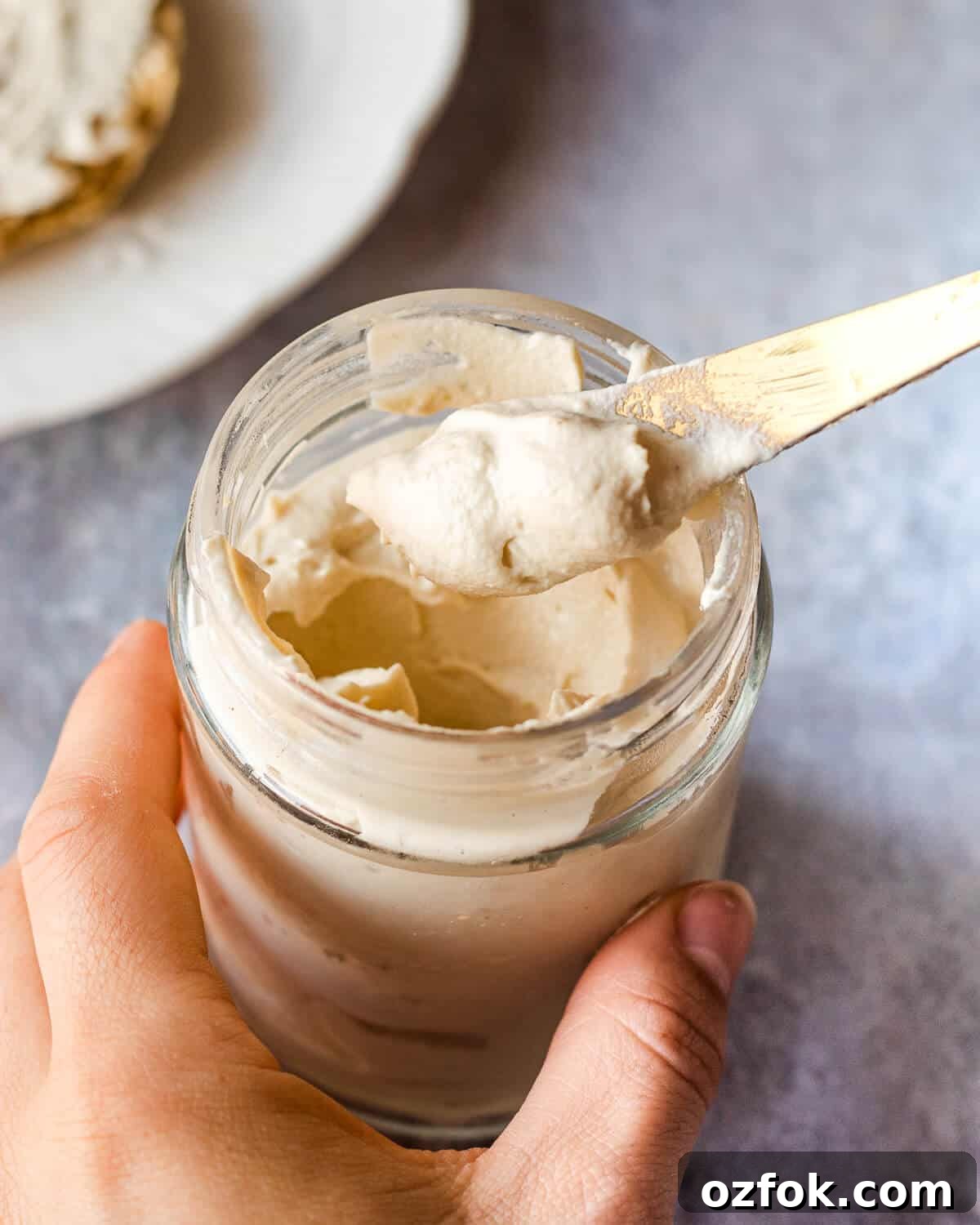 Homemade Vegan Cream Cheese Recipe — Smooth & Tangy Plant-Based Spread 4 A jar of vegan cream cheese. Someone is holding the jar and a knife with some cream cheese on it. There is a plate in the background.