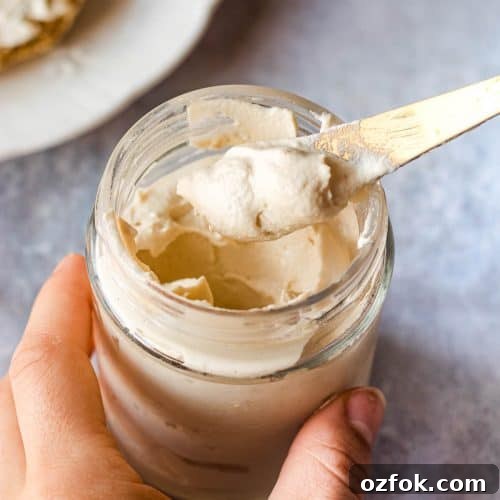 Homemade Vegan Cream Cheese Recipe — Smooth & Tangy Plant-Based Spread 5 A jar of vegan cream cheese. Someone is holding the jar and a knife with some cream cheese on it. There is a plate in the background.