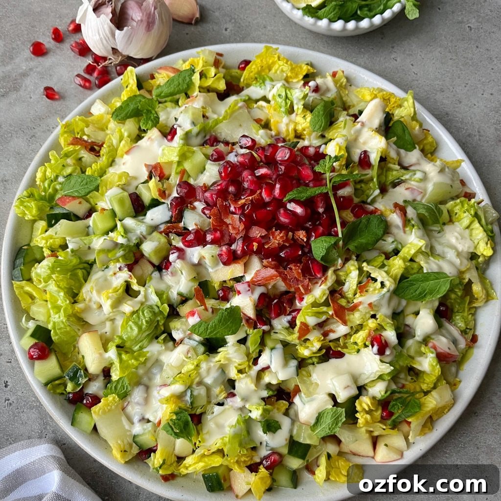 Crisp Prosciutto and Arugula Salad with Parmesan and Lemon Vinaigrette 2 A plate of Crispy Prosciutto Salad topped with pomegranate seeds, chopped cucumber, diced apple, fresh mint leaves, and creamy dressing. Garlic, mint, and a striped napkin surround the plate on a gray surface.
