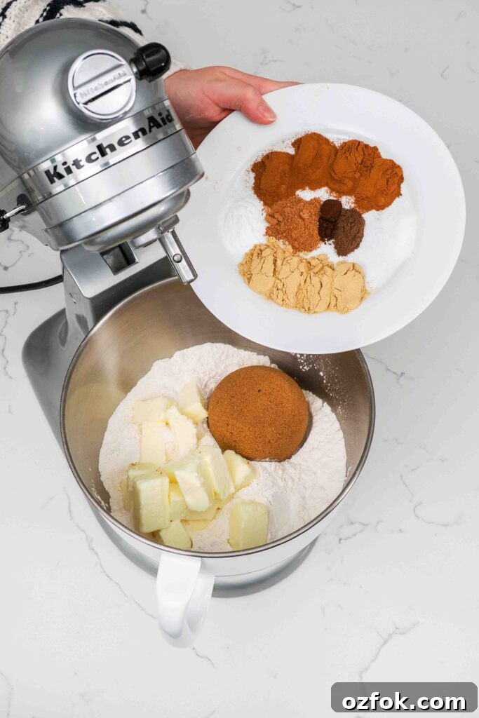 Spices being added to a mixer bowl with flour, brown sugar, and butter.