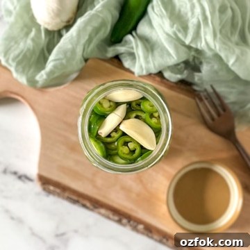 Pickled serrano peppers are shown in a glass jar surrounded by garlic, serrano peppers, and a fork.