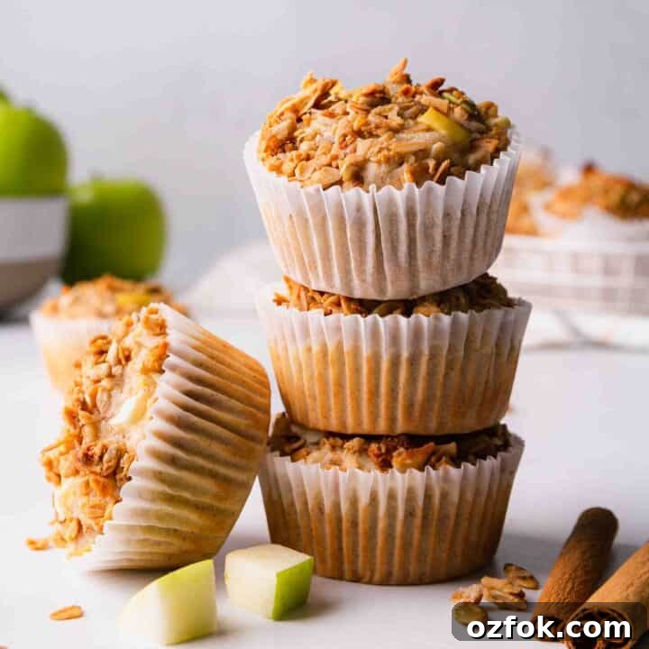 A stack of three gluten-free apple granola muffins with one more leaning on the side of the stack. Surrounded by diced apples and cinnamon sticks.