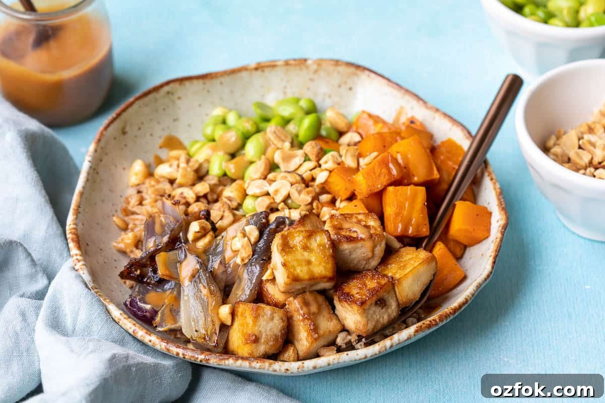 Crispy Tofu Buddha Bowl with Sesame Ginger Dressing 10 crispy tofu buddha bowl with fork next to napkin, jar of peanut sauce, and bowls of edamame and peanuts.