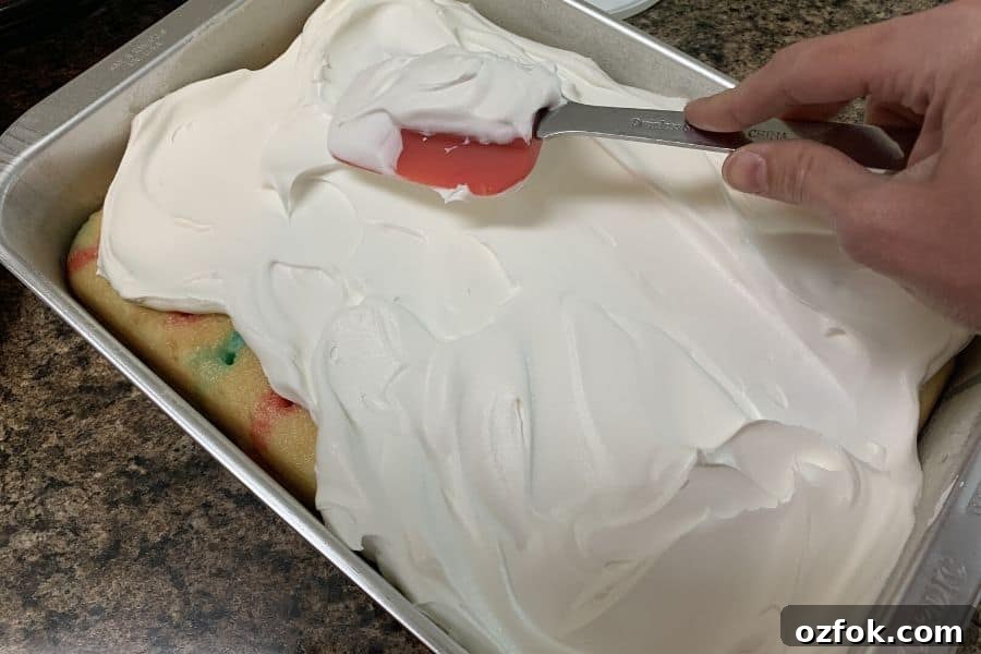 Patriotic Red White & Blue Poke Cake Recipe for 4th of July 9 hand holding a spatula and spreading whipped cream onto a red white and blue poke cake