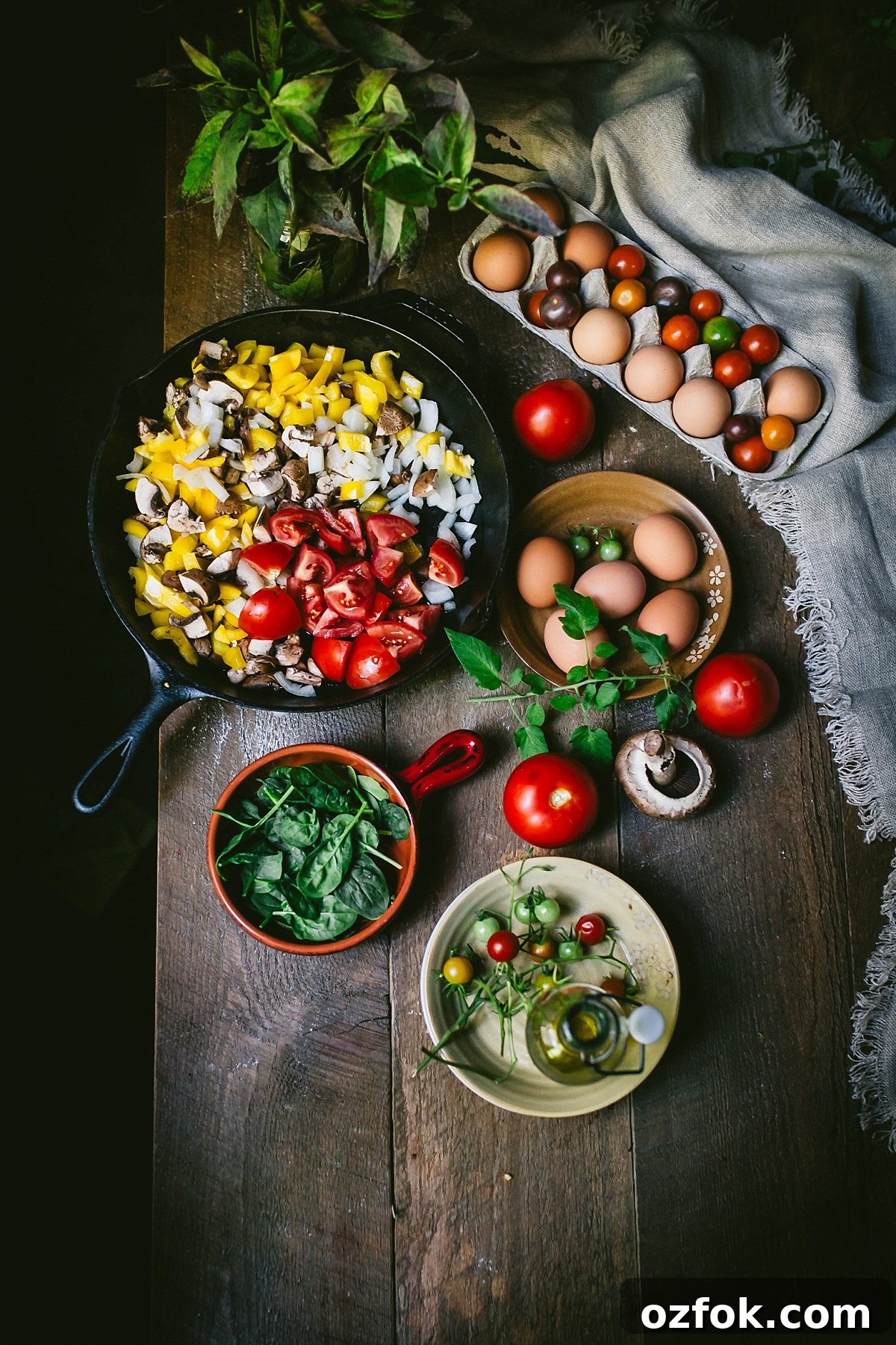 overhead shot of rustic brown table organized with varying bowls overflowing with ingredients for shakshuka: diced peppers, spinach, mushrooms, eggs, and lots and lots of tomatoes