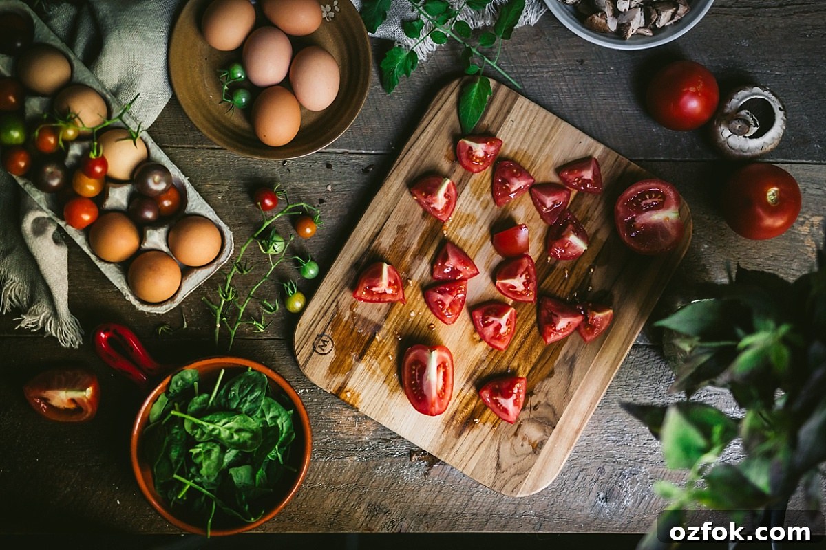 overhead horizontal shot of juicy chopped tomatoes on a cutting board surrounded by basil, mint, spinach, and eggs for shakshuka