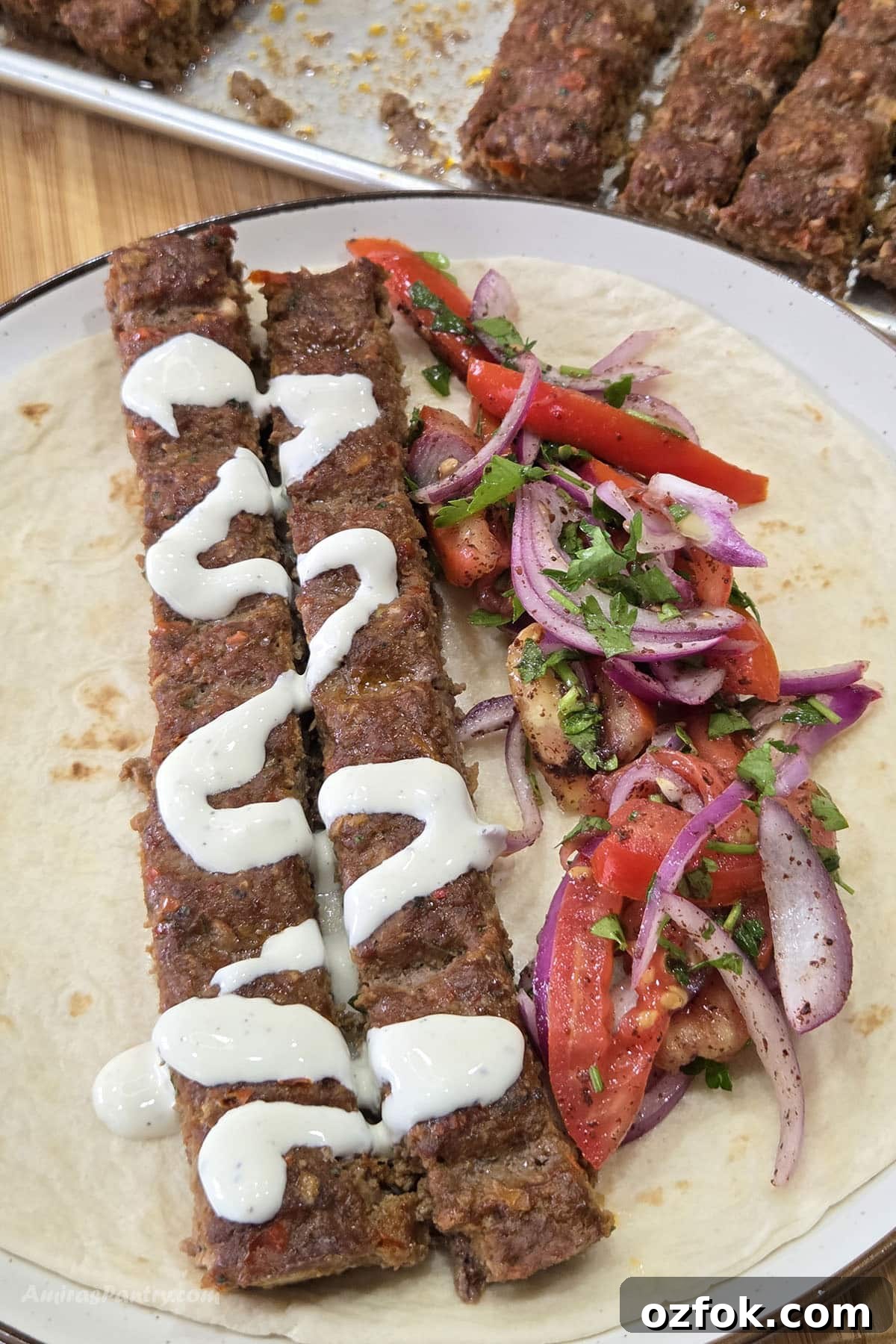 Two grilled Adana kebabs drizzled with creamy garlic sauce are served on a soft flatbread with a fresh tomato, red onion, and parsley salad.
