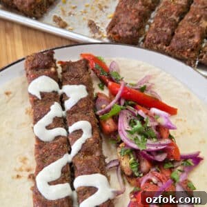 Close-up of two grilled kofta kebabs drizzled with creamy tahini sauce are served on a soft flatbread with a fresh tomato salad. A tray of additional kebabs in the background adds context to this colorful and hearty Middle Eastern-inspired meal.