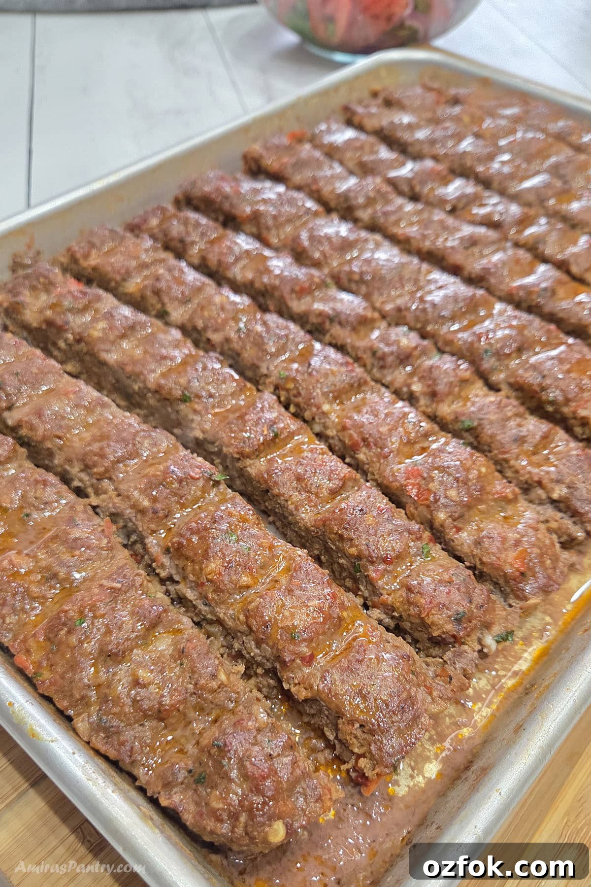 Sheet pan of freshly baked Adana kebabs arranged in neat rows, glistening with flavorful juices and rendered fat. The golden-brown ground meat kebabs are scored into portions and ready to serve, with a bowl of fresh salad blurred in the background.