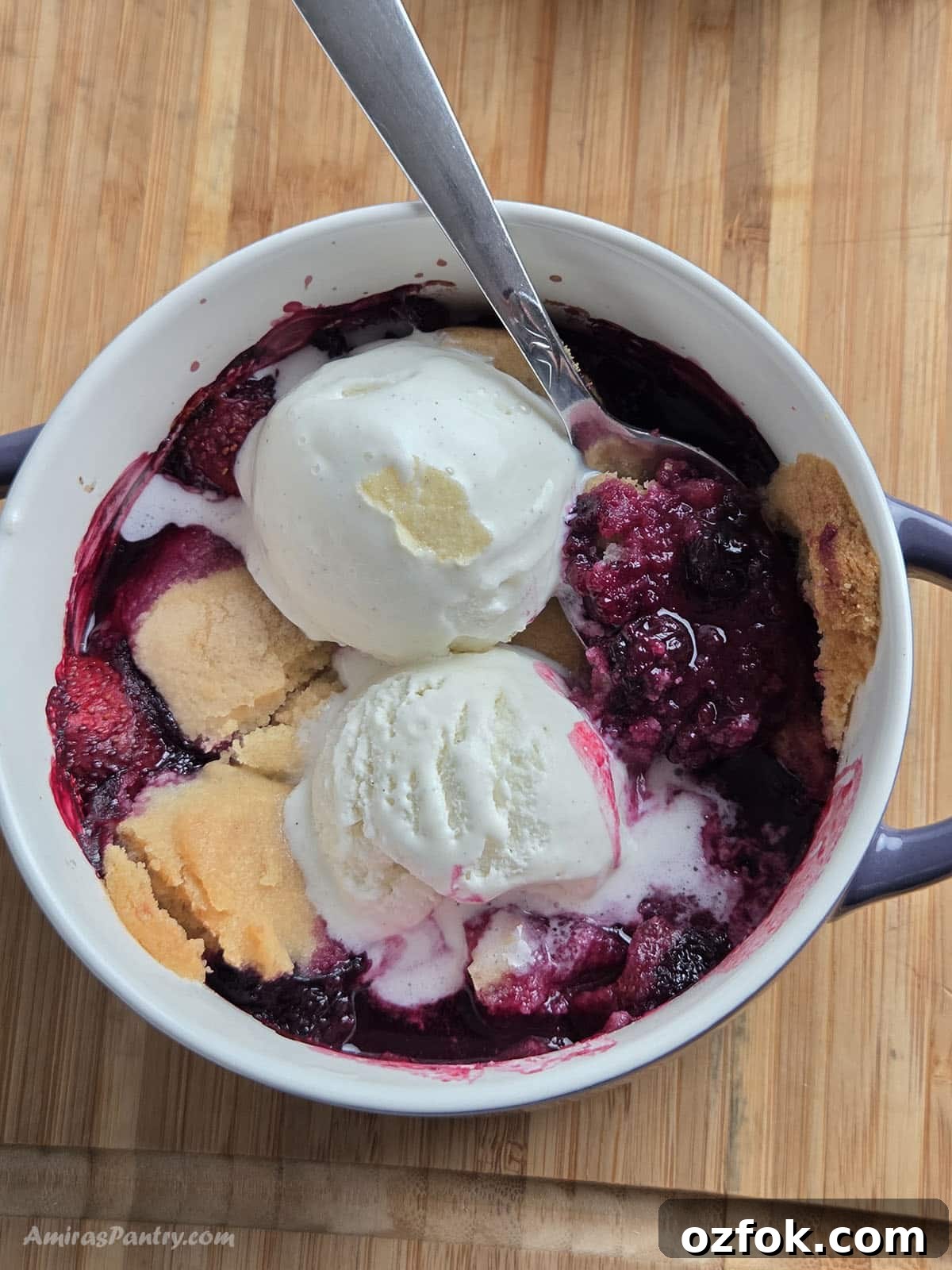 Overhead view of a two-ingredient berry cobbler in a small dish, topped with two scoops of melting vanilla ice cream and a spoon, set on a wooden table.