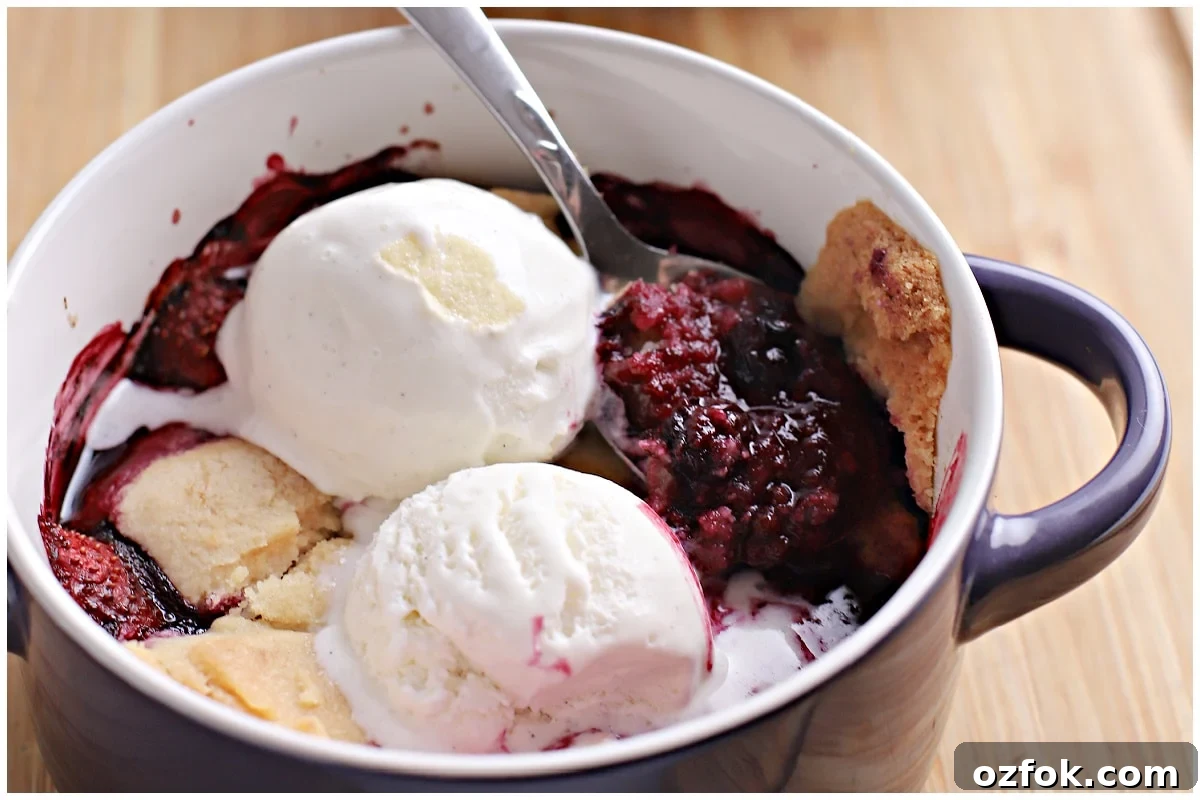 Close-up angled shot of baked berry cobbler with golden biscuit topping in a blue handled dish, topped with two melting scoops of vanilla ice cream and a spoon.