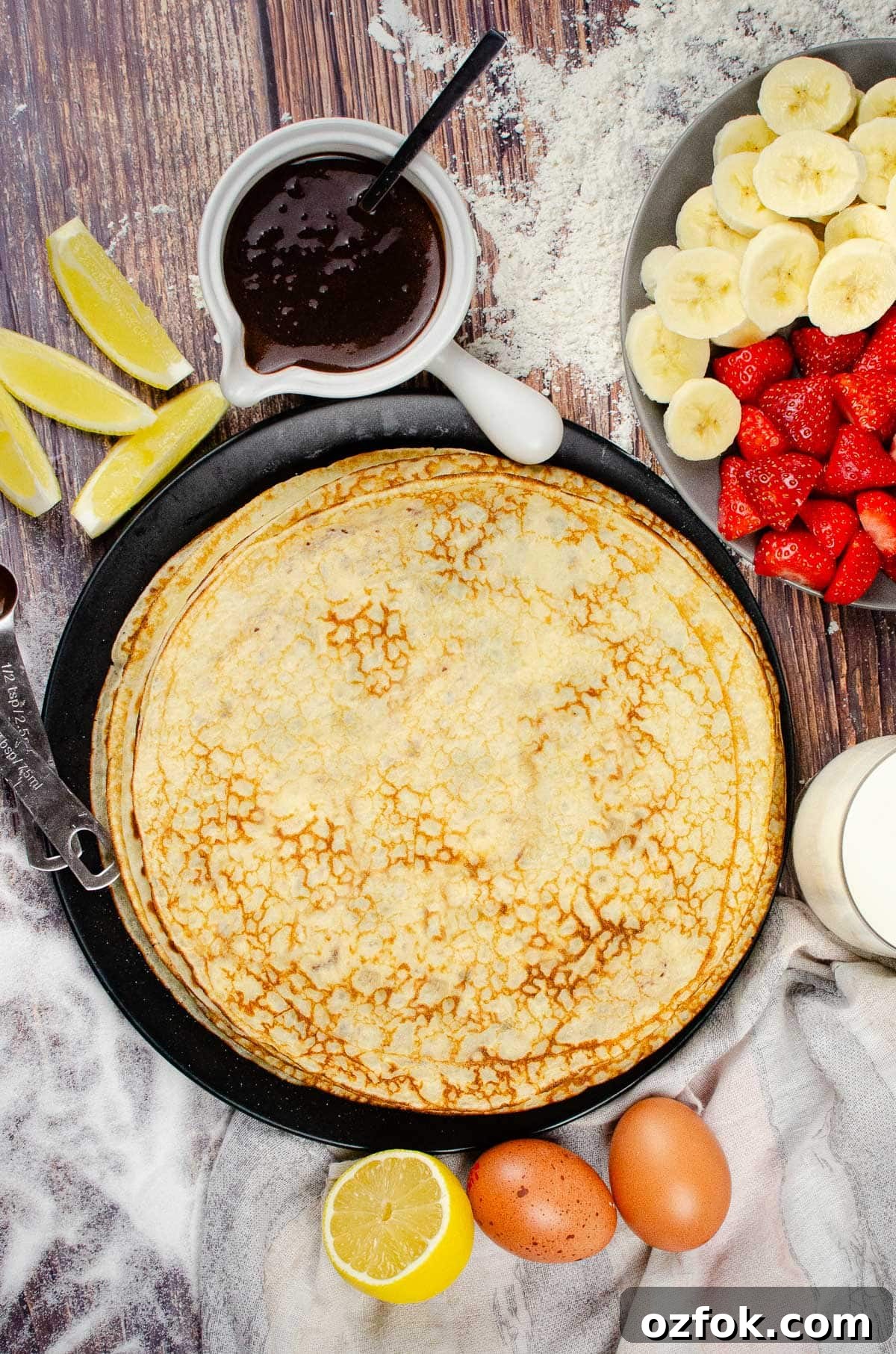 A stack of Easy English Pancakes on a black plate on a surface of scattered sugar with a white pot of chocolate sauce, plate of chopped strawberries and bananas, a glass of milk, two eggs, slices of lemon,½ tablespoon of vegetable oil and ½ teaspoon of vanilla extract on the side